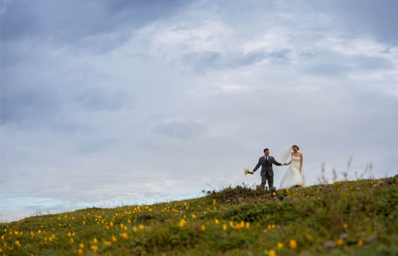 A bride and groom walking on a hillside of yellow flowers in Bodega Bay overlooking the Pacific Ocean.